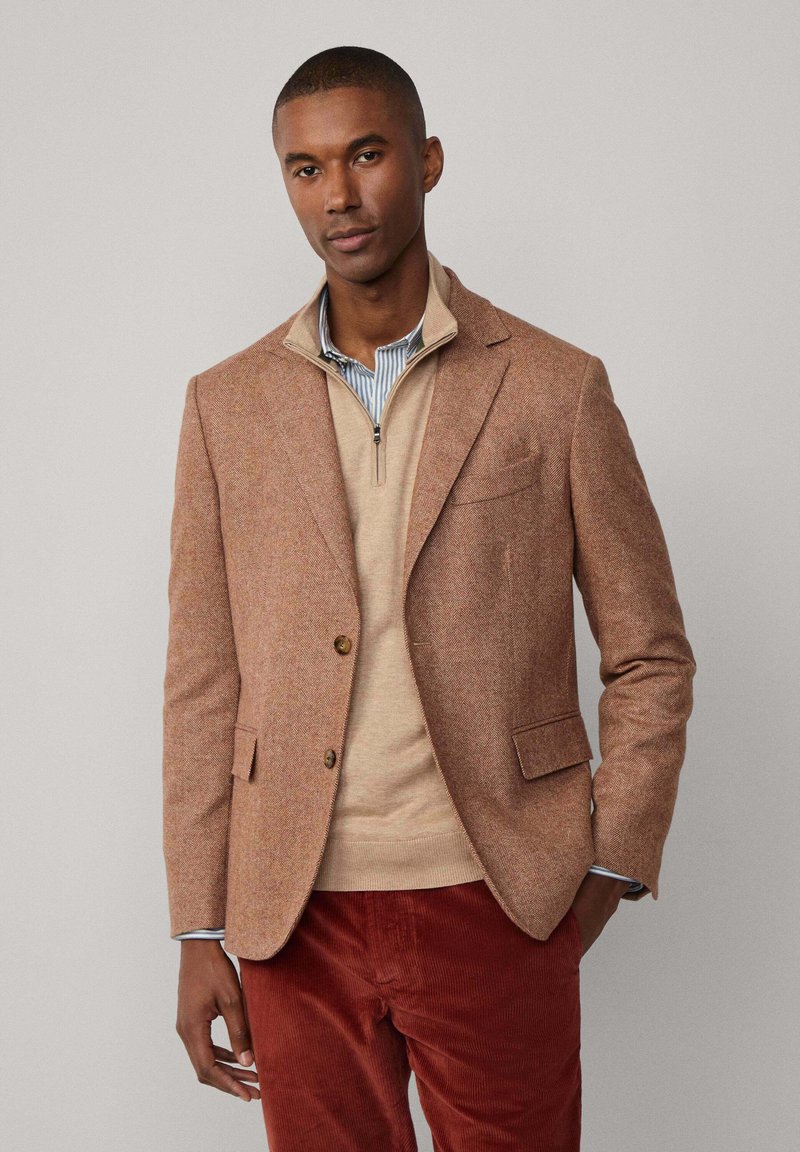 Bruine visgraat blazer van gestructureerde stof, gelaagd over een beige rits trui. Past goed bij rode corduroy broek met een subtiel patroon.