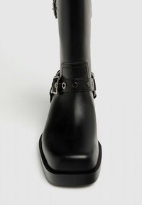 Bota de cuero negro hasta la rodilla con punta cuadrada, que presenta una correa con herrajes plateados y studs circulares, acabado mate y suela robusta.