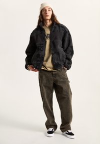 Schwarze Fleecejacke mit strukturiertem Stoff und schwarzen Einsätzen, kombiniert mit einem beigen T-Shirt und dunklen, lockeren Hosen. Schwarz-weiße Sneakers.