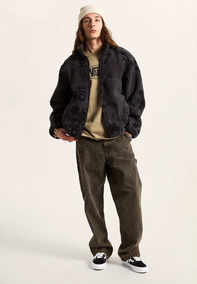 Schwarze Fleecejacke mit strukturiertem Stoff und schwarzen Einsätzen, kombiniert mit einem beigen T-Shirt und dunklen, lockeren Hosen. Schwarz-weiße Sneakers.