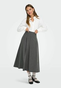 Falda larga de cuadros grises con pliegues, combinada con una camisa blanca de botones y calcetas de encaje. Acentuada por unos zapatos negros de estilo Mary Jane.