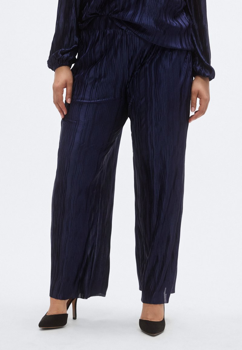 Meresinine laia lõikega plisseeritud püksid siidja kangaga. Omavad siledat tekstuuri ja elastset vööd; kombineeritud mustade kõrgete kontsadega kingadega.