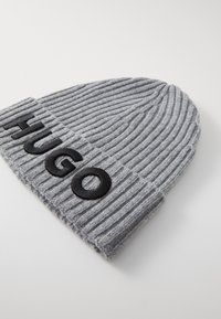 Beanie gri tricotat cu textură înrădăcinată și o ecuson din piele neagră pe care este afișat "HUGO" pe manseta pliată.