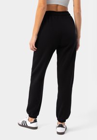 Melni jogginga bikses ar elastīgu jostasvietu, sašaurinātiem kauliem un mīkstu tekstūru. Valkātas ar baltām sporta kurpēm, kurām ir melnas svītras.