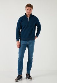 Sudadera azul marino con cuello de cremallera y mangas largas, combinada con jeans de un azul claro y zapatillas oscuras con detalles en gris. Textura suave y ajuste casual.