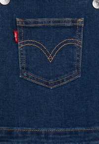 Džínové kombinézy mají tmavě modrou látku s klasickým designem zadní kapsy. Zlaté prošívání vytváří charakteristický obloukový vzor nad kapsou.