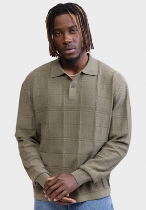 Νεαρός άνδρας με dreadlocks μέχρι τους ώμους, φοράει μακρυμάνικο πουλόβερ πόλο χακί με τρύπες και μπλε τζιν, με τα χέρια σταυρωμένα.