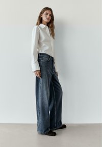 Bílá košile na knoflíky spojená s tmavě modrými širokými džínami s volným střihem a jemnými šicími detaily. Jednoduchá obuv doplňuje vzhled.