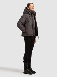 Szara puchowa kurtka z kapturem, z zamkiem błyskawicznym z przodu i skośną kieszenią na zamek. Modelka ma na sobie czarne legginsy i kozaki za kolano.