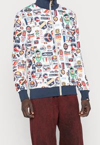 Vit zip-up jacka med många färgglada lappar och logotyper, med marinblå krage och muddar. Matchas med mörkbruna byxor.