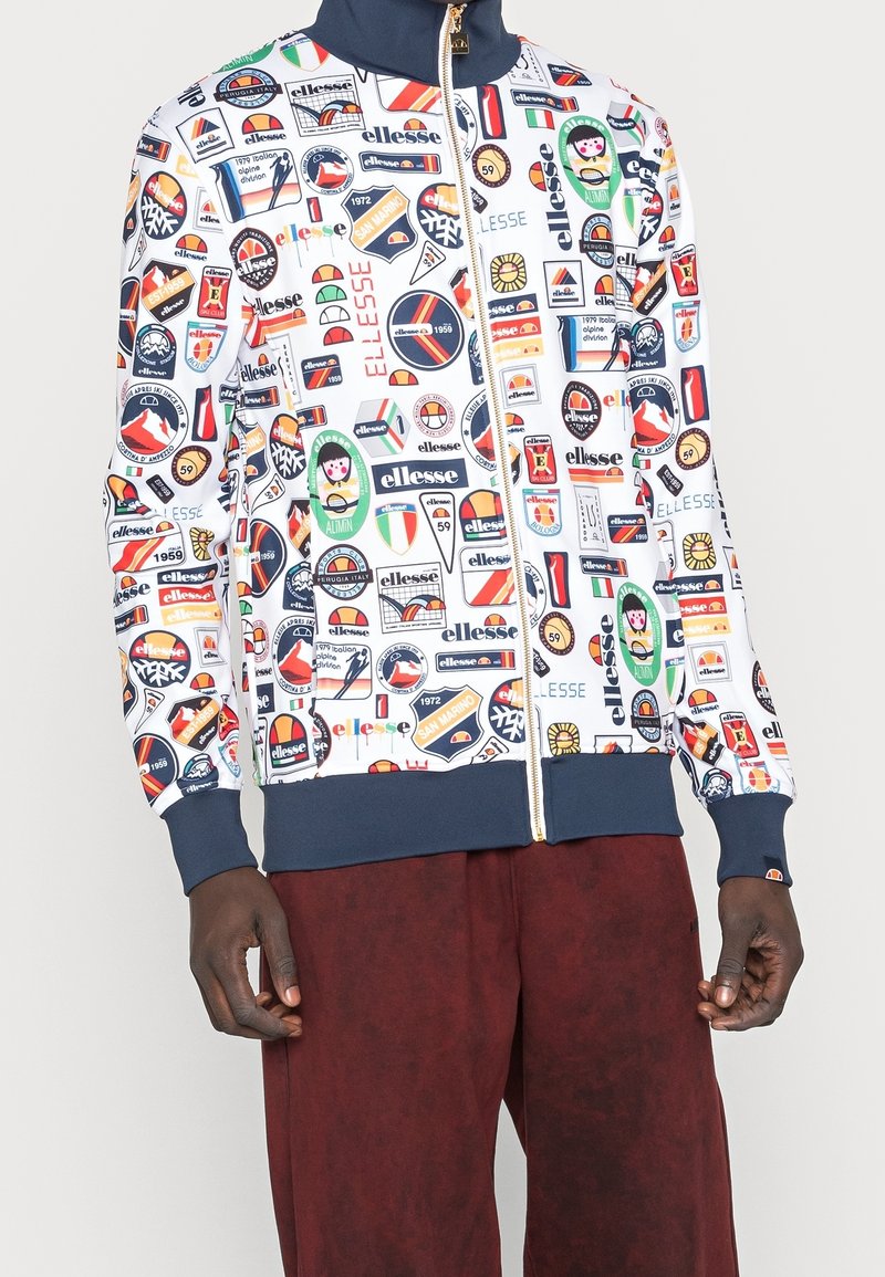 Vit zip-up jacka med många färgglada lappar och logotyper, med marinblå krage och muddar. Matchas med mörkbruna byxor.