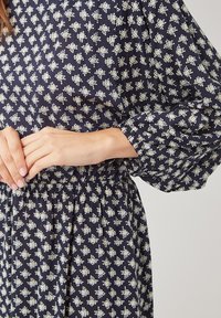 Mujer vestida con un vestido azul marino con un patrón geométrico blanco, que presenta mangas largas abombadas y una cintura elástica, con las manos suavemente entrelazadas a la altura de la cintura.