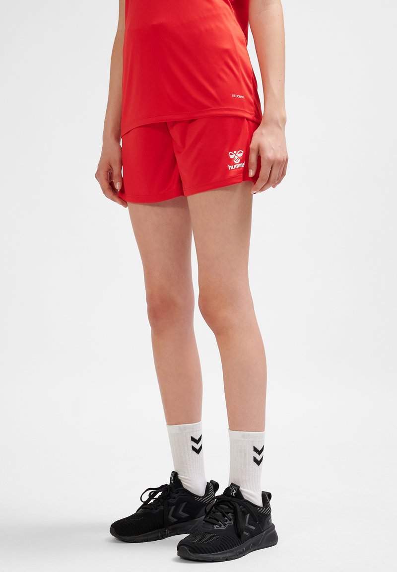 Röda sportshorts i lätt tyg med en logotyp på sidan, kombinerade med svarta sneakers och vita strumpor med svarta vinklar.