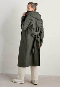 Olijfgroene trenchcoat met capuchon, ceintuur aan de achterkant en knoopdetails. Gecombineerd met crèmekleurige broek en grijze sneakers.