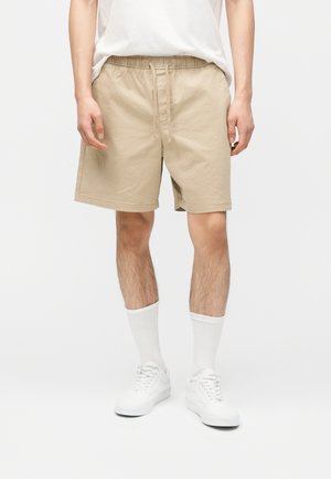 Hombre con pantalones cortos beige con cordón, calcetines blancos hasta el tobillo, zapatillas blancas y camiseta blanca metida, de pie contra un fondo liso.