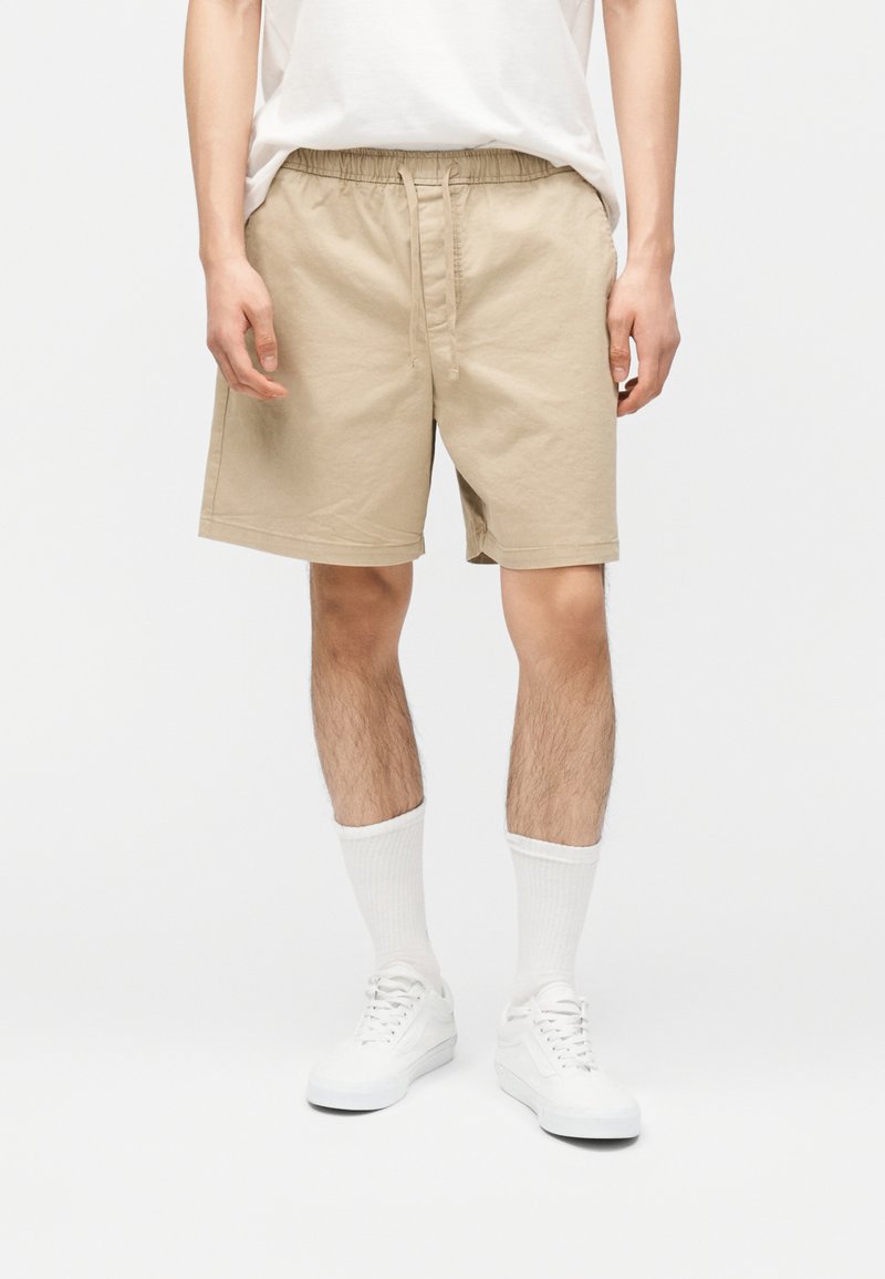 Άνδρας που φοράει μπεζ σορτς με κορδόνι στη μέση, άσπρες κάλτσες μέχρι τον αστράγαλο, λευκά αθλητικά παπούτσια και λευκό μπλουζάκι μακρυμάνικο, κρυμμένο μέσα στο σορτς, στέκεται μπροστά σε απλό φόντο.