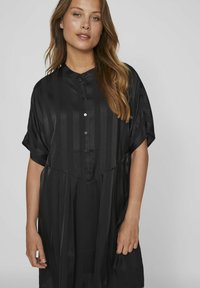 Abito camicia nero con maniche corte, chiusura a bottoni, righe verticali e una vestibilità rilassata. Il tessuto appare liscio e leggermente luminoso.