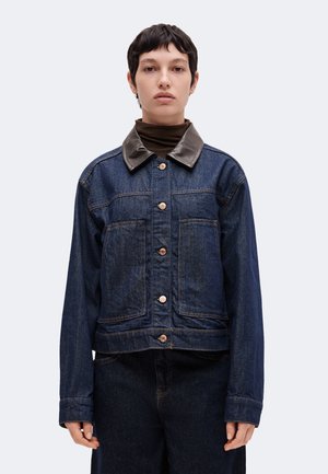 Personne aux cheveux courts et foncés portant une veste en denim foncé avec de grandes poches avant et un col en cuir noir sur un col roulé marron.
