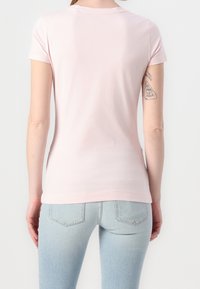 Kobieta w jasnoróżowej koszulce z krótkim rękawem i jasnoniebieskich dżinsach, pokazana od tyłu, z widocznymi tatuażami na lewym ramieniu.