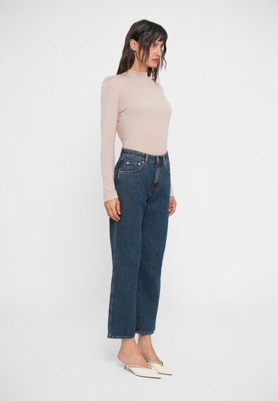 Top beige claro de manga larga combinado con jeans oscuros de talle alto y corte holgado. La modelo lleva zapatos blancos de tacón bajo. Fondo liso.