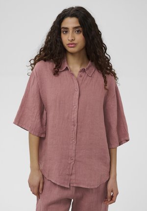 Femme aux longs cheveux bouclés portant une chemise ample rose poussière à boutons et un pantalon assorti, debout devant un fond clair uni.