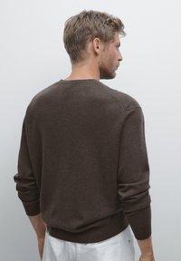 Uomo con capelli corti e castani, che indossa un maglione marrone scuro e pantaloni bianchi, leggermente girato a destra su uno sfondo semplice.