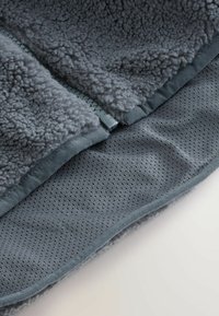 Szary polarowy materiał o miękkiej, puszystej fakturze nakłada się na gładką, oddychającą siatkową warstwę. Posiada zamek wzdłuż jednej krawędzi.