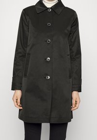 Svart knappad kappa med krage, långa ärmar, sidofickor och sydda detaljer på ärmslut och nederkant, bärs över en vit turtleneck och svarta byxor.