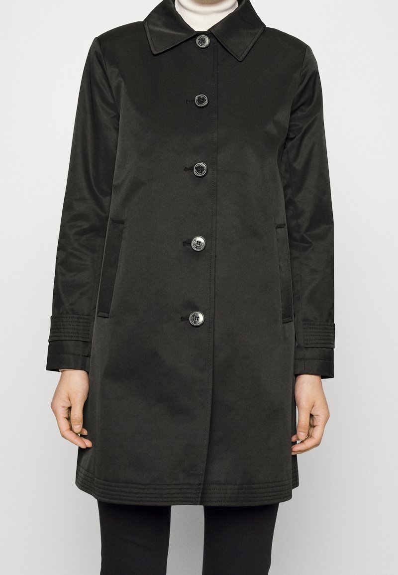 Svart knappad kappa med krage, långa ärmar, sidofickor och sydda detaljer på ärmslut och nederkant, bärs över en vit turtleneck och svarta byxor.