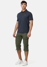 Marineblå polo med korte ærmer og et orange logo, parret med grønne cargo-shorts med flere lommer. Båret med hvide sneakers.
