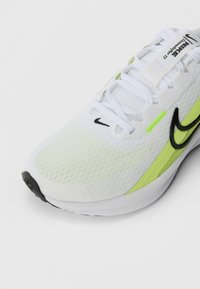Hvidt atletiksko med en åndbar mesh-overdel, sort Nike-swoosh og neongrønne accenter på siden og hælen. Hvide snørebånd og struktureret sål.
