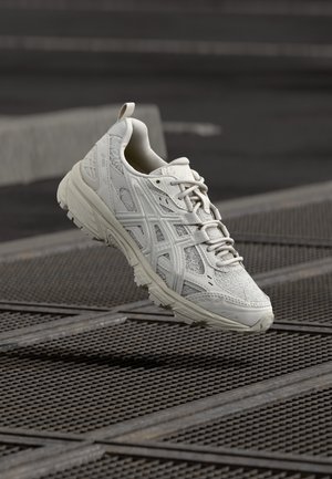 Biały but do biegania Asics zawieszony nad metalową kratą, ukazujący szczegółową fakturę i elementy designu.