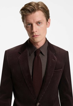 Bruin corduroy blazer met subtiele ribbels, gecombineerd met een donkerbruine overhemd en bijpassende gebreide stropdas. Bevat een pochet detail.