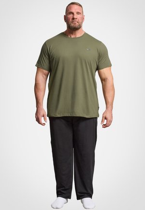 Muskuløs mand står med fronten mod kameraet iført en olivengrøn kortærmet t-shirt, sorte bukser og hvide sokker mod en ensfarvet baggrund.