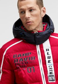 Red puffer jacket with a navy hood, featuring embroidered logos and white accents. Puff quilted texture and snap-button closure details.