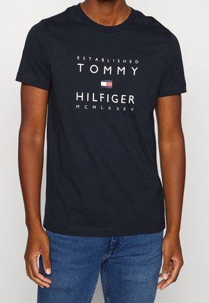 Schwarzes Baumwoll-T-Shirt mit weißem Text "ESTABLISHED TOMMY HILFIGER" sowie einer Logo-Flagge und der Jahreszahl "MCMLXXXV" darunter. Kurze Ärmel, Rundhalsausschnitt.