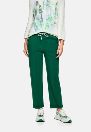 Person wearing green drawstring pants, white sneakers with green accents, and a white long-sleeve top with abstract green and blue print.