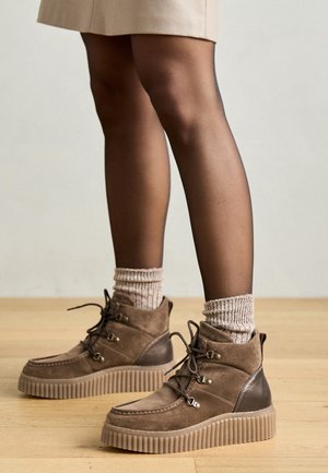 Beine, die durchsichtige schwarze Strumpfhosen, beige gerippte Socken und braune Wildleder-Schnürstiefel mit Plateau auf hellem Holzfußboden tragen.