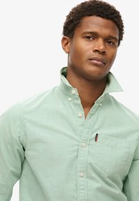 Camisa de botonadura verde claro hecha de una tela texturizada, con cuello, un bolsillo en el pecho y un pequeño logo encima del bolsillo.