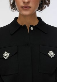Giacca nera con collo e due tasche frontali, caratterizzata da una chiusura con bottoni argentati e decorazioni con spille gioiello su ogni tasca.