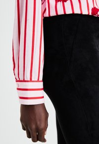 Manche de chemise en coton rayée rouge et blanche avec poignet à bouton, associée à un pantalon noir d'apparence suédée. Accent sur la texture et le contraste des couleurs.