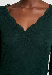 Mörkgrön spetsblus med en vågig V-ringning och lång ärm. Det intrikata blommönstret ger struktur och detaljer.