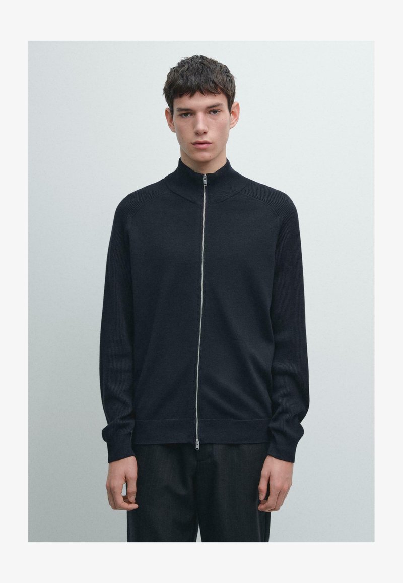 Marineblauer Zip-Up-Pullover mit hohem Kragen, gerippter Textur an Schultern und Bündchen sowie glattem Körper, kombiniert mit dunkler Hose.