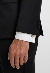Black tailored suit jacket with a white cuff, featuring a silver square cufflink. Subtle texture on the fabric of the jacket and cuff.