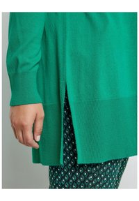 Zelený pletený svetr s žebrovanou strukturou a bočním rozparkem. Kombinovaný s tmavě zelenými kalhotami s geometrickým vzorem.