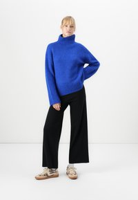 Frau steht mit einer Hand auf der Hüfte, tragen einen leuchtend blauen Rollkragenpullover, weite schwarze Hose und Sneaker mit Leopardenmuster.