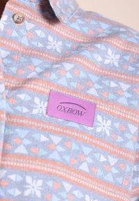 Chemise à boutons avec un motif géométrique et floral pastel en bleu, orange et blanc, avec une étiquette rose portant le texte "OXBOW".