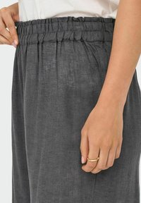 Šedé lněné kalhoty s nařaseným elastickým pasem, mají měkkou texturu a volný střih. Ruka částečně zobrazená, jak drží zlatý prsten.