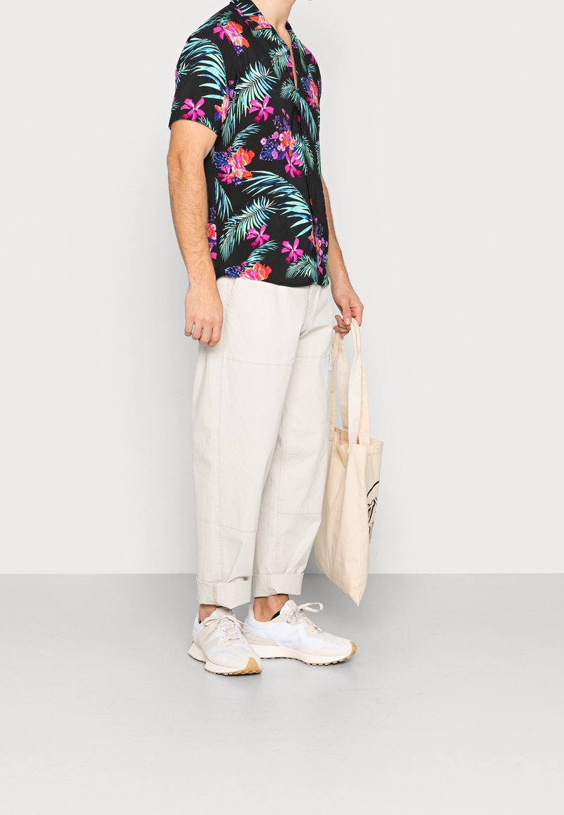 Short-sleeve black shirt with colorful floral and leaf print, paired with light beige, loose-fitting pants and white sneakers. Holding a tote bag.