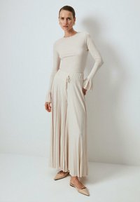 Žena oblečená v dlhom béžovom tričku s volánovými manžetami, širokých plisovaných béžových nohaviciach a béžových špicatých balerínach stojí pred jednoduchou stenou.